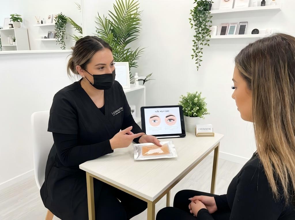 A professional LASHMAITRE technician consulting with a client, explaining safe methods to treat eye irritations and showing how to remove a stye without damaging lash extensions using a warm compress.