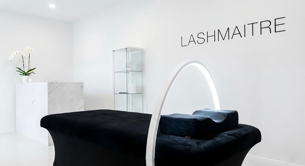 Interior of a luxury lash studio featuring a sleek white Half Moon eyelash extension light arching over a black velvet lash bed, casting a bright, shadowless glow. LASHMAITRE professional equipment.