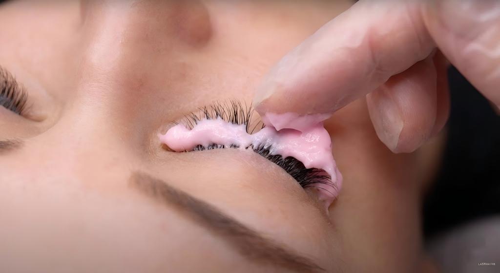 Close-up demonstration showing a professional technician applying a thick layer of LASHMAITRE pink cream remover precisely to the base of the extensions, illustrating the safe method of how to dissolve eyelash extension glue without skin contact.