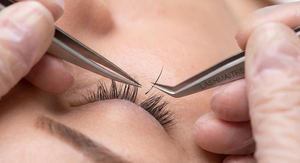 High-resolution macro photography showing a professional lash artist using LASHMAITRE tweezers to isolate a natural lash and precisely apply a single extension. This demonstrates the 1:1 technique used for creating classic natural eyelash extensions.