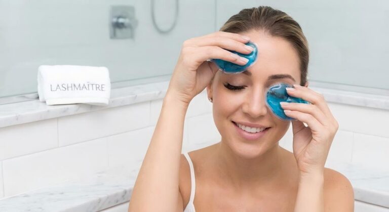 A woman with eyelash extensions applying blue gel cold compresses to her eyes in a bathroom, demonstrating one of the most effective at-home eyelash extension allergic reaction remedies to reduce puffy eyelids.