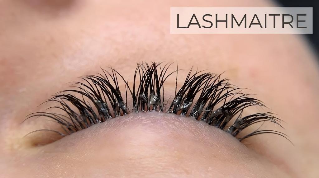 A LASHMAITRE educational macro photograph showing a close-up example of bad eyelash extensions, characterized by visible adhesive clumps, stuck-together lashes, and uneven spacing on a closed eye.