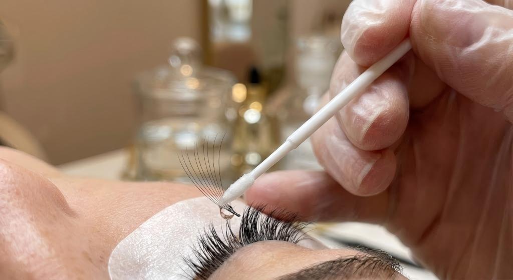 Macro photography showing the precise application of LASHMAITRE eyelash extension bonder using a micro-swab to the base of a volume fan to instantly cure the adhesive.