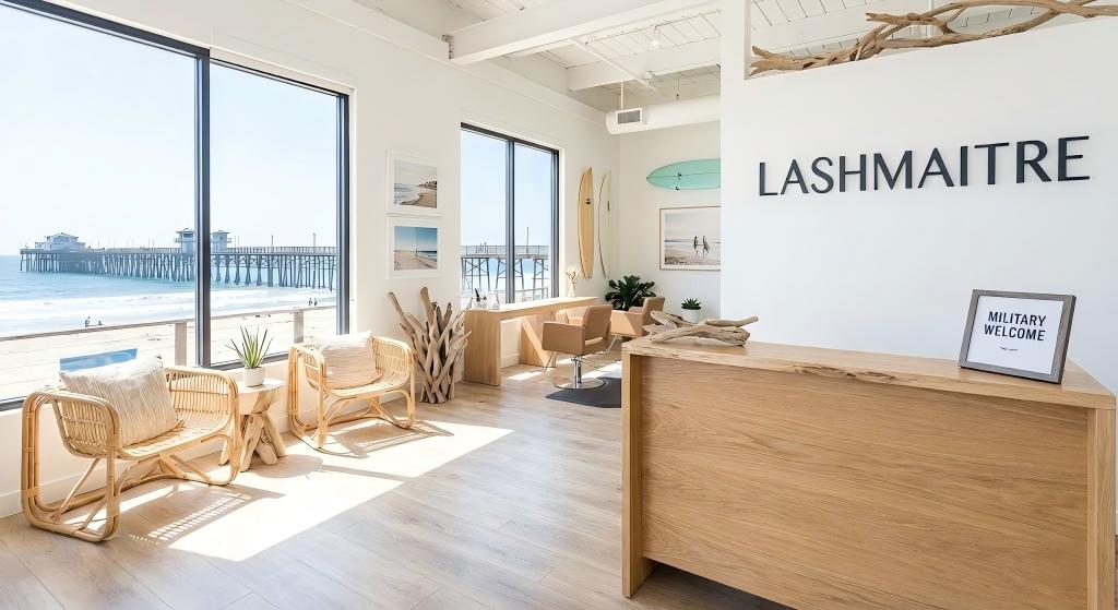 The interior of LASHMAITRE, a salon specializing in eyelash extensions Oceanside, featuring bright beach decor, a "Military Welcome" sign on the reception desk, and large windows overlooking the Oceanside Pier and ocean.