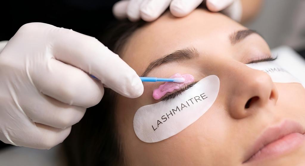 Close-up technical shot of a professional lash artist using a micro-swab to precisely apply thick, pink LASHMAITRE cream remover for eyelash extensions to the base of a client's closed lashes, demonstrating safe removal technique.