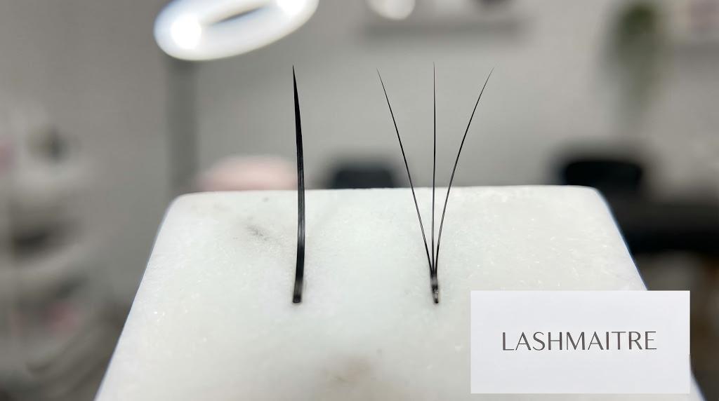 A macro close-up photograph comparing LASHMAITRE eyelash extension thicknesses, showing a single thick classic lash next to a fan of fine volume lashes on a marble tile under professional lighting.