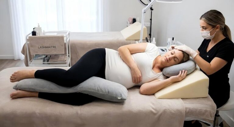 A pregnant client at LASHMAITRE comfortably positioned on her side with supportive body and wedge pillows during a professional appointment, demonstrating safe maternity lash service protocols.