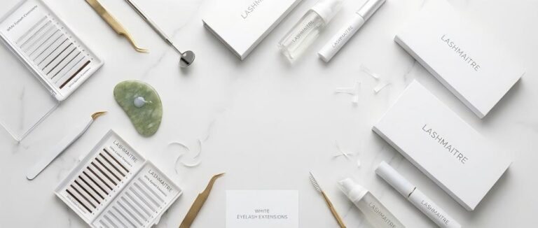 Flat lay of LASHMAITRE white eyelash extensions trays, gold and silver tweezers, lash mirror, adhesive on a jade stone, and branded packaging on a white marble surface.