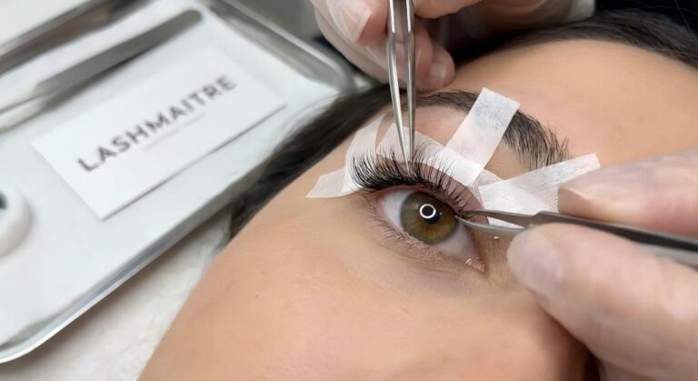 "Close-up view of a LASHMAITRE stylist performing a specialized taping technique for correcting round eye shape with lash extensions, using tape to isolate outer corner lashes for precise application."