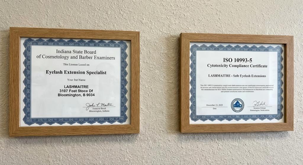 Two framed documents on a textured salon wall at LASHMAITRE in Bloomington, Indiana. The left frame holds an 'Indiana State Board of Cosmetology' license for an 'Eyelash Extension Specialist'. The right frame holds an 'ISO 10993-5 Cytotoxicity Compliance Certificate' for 'LASHMAITRE - Safe Eyelash Extensions'.