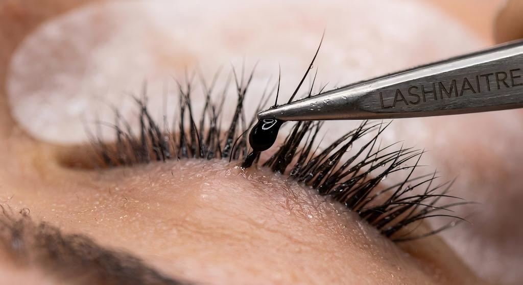 Macro photography of a black eyelash extension adhesive droplet on a lash, showing a smooth, glossy finish in high humidity, resisting shock polymerization with the LASHMAITRE logo engraved on the tweezers.